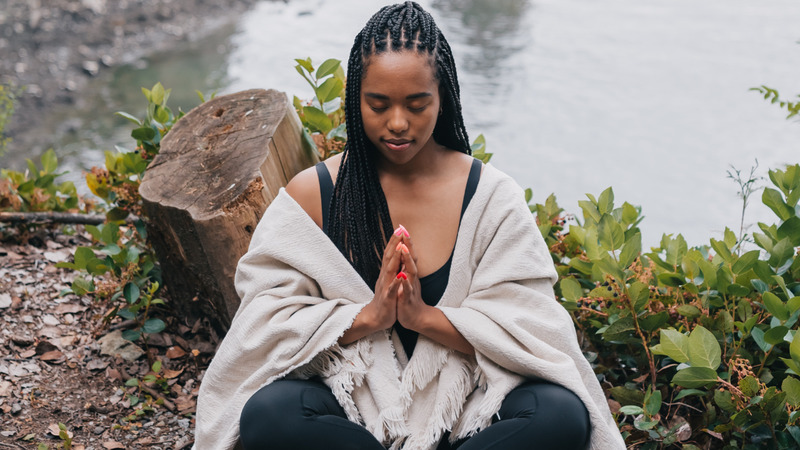 Person with braided hair sitting cross-legged near water, hands in prayer position, in an outdoor setting with plants and a tree stump.